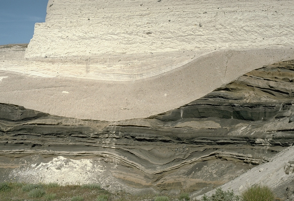 This outcrop shows light-colored deposits from the 3,500-year-old Minoan eruption of Santorini filling a valley that eroded into darker tephra layers of Pleistocene age. The lower beige-colored unit filling the valley is a pumice-fall deposit from early in the eruption. It is overlain by laminated pyroclastic surge deposits that were produced when water came into contact with the magma reservoir as the volcano collapsed into the sea. The upper lighter-colored layer truncating both these deposits is a pyroclastic flow deposit. Photo by Lee Siebert, 1994 (Smithsonian Institution).