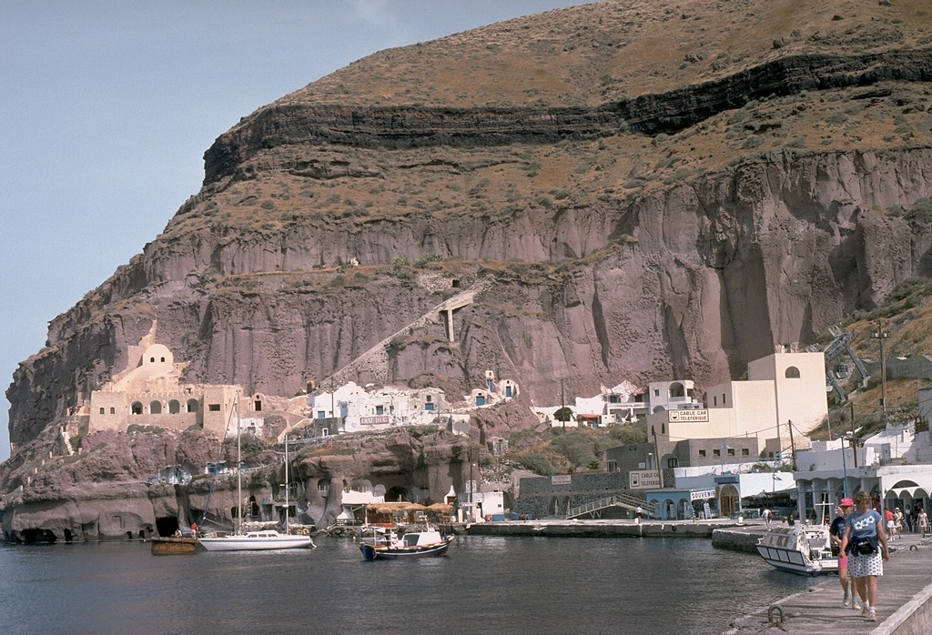 The headland of Cape Thera above Phira harbor. The light-brown, cliff-forming unit above the harbor is the Cape Thera Ignimbrite. Above the cliffs is a sequence of sloping pyroclastic deposits and paleosols, which underlies the prominent thinner dark-colored layer at the top of the photo. This is the Middle Pumice unit, a welded Plinian pumice-fall deposit erupted about 100,000 years ago and thought to originate from a vent west of Phira. The lower-angle slopes above it are lithic breccias of the Middle Pumice eruption. Photo by Lee Siebert, 1994 (Smithsonian Institution).