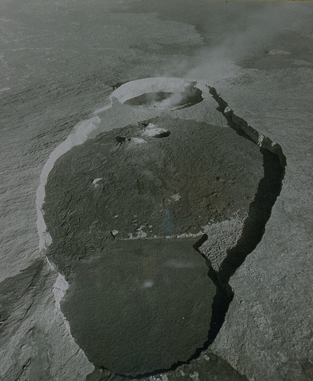 An aerial view looks NW across the axis of the elongated summit crater of Erta Ale volcano.  The summit depression is formed by several coalescing craters.  Several steep-walled pit craters cut the floor of the elliptical crater.  At the time of this 1971 photo, steam rises from the northern of two active lava lakes.  Lava overflows from the smaller southern pit crater (near the top of the area covered by dark lava flows) spread across much of the broad crater floor and spilled over its SE rim at the bottom of the photo. From the collection of Maurice and Katia Krafft.