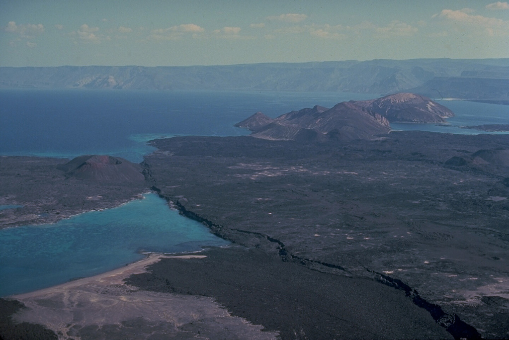 The Ardoukôba (Asal) Rift in Djibouti, trending NW from the Red Sea, contains a broad area of youthful fissure vents between Lake Asal and the Ghoubbat al Kharab gulf.  This view looks from the north towards the gulf.  Regional faults of the Ardoukôba Rift trending NW-SE cut lava-flow fields.  A cinder cone appears at the left, and a group of tuff cones lie at the upper right.  The youngest lava flows cover lake sediments deposited 5300 years ago.  An eruption in 1978 produced a small cinder cone and lava flows. Copyrighted photo by Katia and Maurice Krafft, 1976.