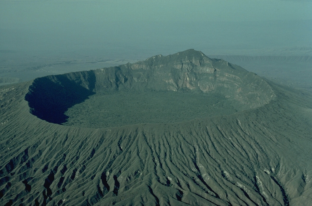 The central cone of Longonot volcano is truncated by a small steep-walled caldera that is 1.8-km wide.  Erosionally furrowed ash deposits related to the caldera-forming eruptions mantle its slopes.  Trachytic lava flows up to 30-m thick that built the central cone can be seen in the far west caldera wall.  The caldera floor is covered by post-collapse aa lava flows .  A line of small craters extends from the SW wall to the center. Copyrighted photo by Katia and Maurice Krafft, 1976.
