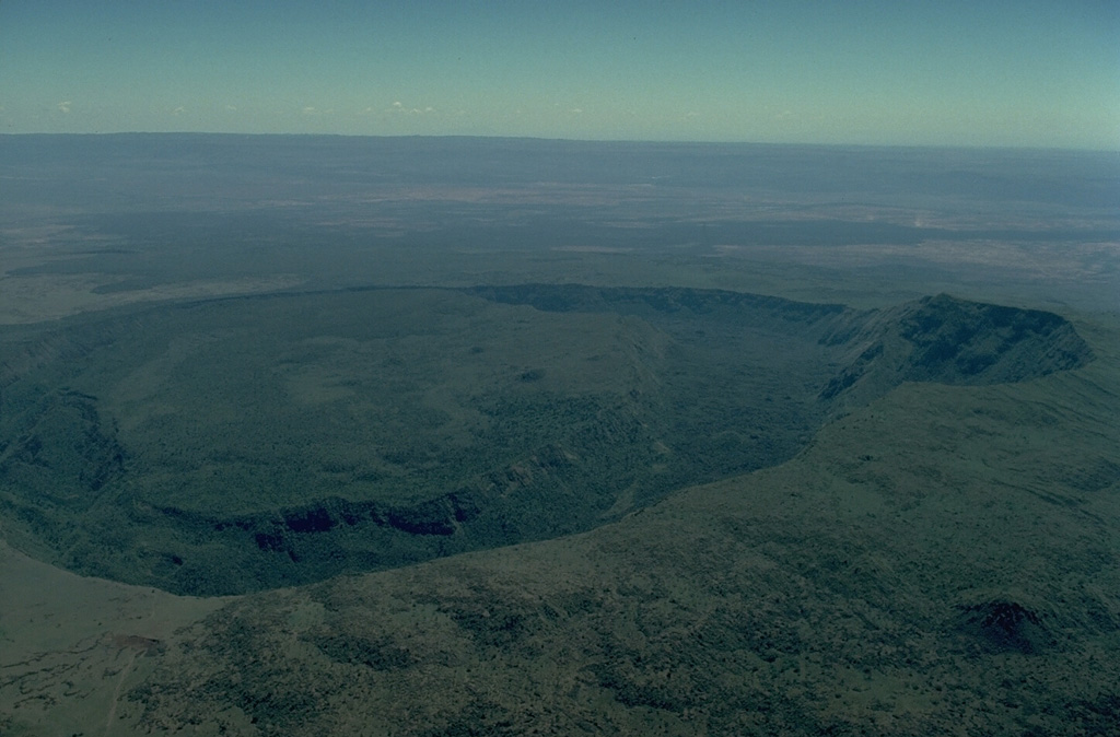 Suswa is a broad low shield volcano that is cut by the southernmost caldera of the Kenya rift.  The 8 x 12 km caldera is filled by a broad lava cone.  An unusual 5-km-wide ring graben, seen here from the west, cuts the lava cone.  The ring graben isolates a tilted island block (left).  Ol Doinyo Nyukie ("The Red Mountain") forms the summit of Suswa volcano (right).  The latest eruptions of Suswa have originated from parasitic vents that have issued still-unvegetated lava flows that may be only a century or so old. Copyrighted photo by Katia and Maurice Krafft, 1976.