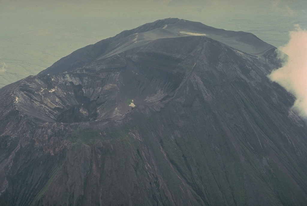 This 1977 photo from the NW shows the summit crater of Ol Doinyo Lengai volcano prior to a decade-long eruption that began in 1983.  The northern crater (left) has been active during historical time.  An older southern crater is located behind the summit, which is the dark, rounded ridge below the upper right horizon.  Prior to 1983 the northern crater was deep, with a steep-walled inner pit.  Beginning in 1983, the slow effusion of lava flows on the crater floor began filling it. Copyrighted photo by Katia and Maurice Krafft, 1977.
