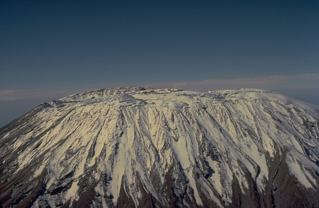 Massive Kilimanjaro, Africa's highest mountain, consists of three very large stratovolcanoes constructed along a NW-SE trend.  Seen here from the SW, the ice-capped, 5895-m-high summit is cut by a 2.4 x 3.6 km caldera that gives the summit of the central stratovolcano (Kibo), an elongated broad profile.  Most of Kilimanjaro was constructed during the Pleistocene, but a group of youthful-looking nested summit craters are of apparent Holocene age.  Fumarolic activity continues at several locations on the summit plateau. Copyrighted photo by Katia and Maurice Krafft, 1977.