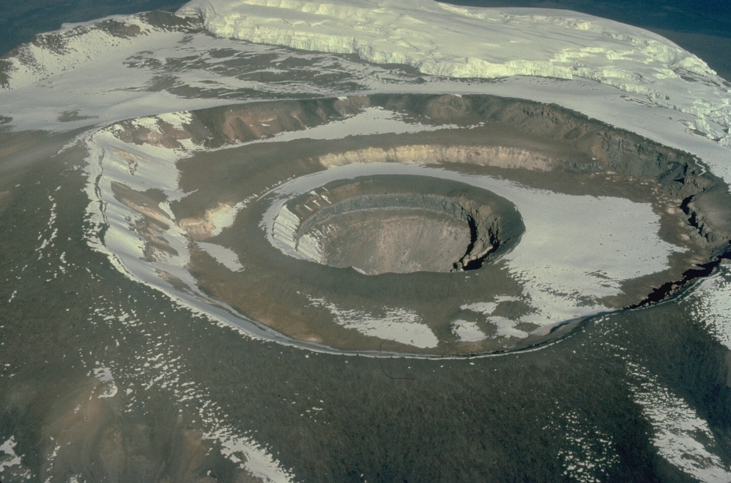 An aerial view from the SE overlooks three nested craters at the summit plateau of Tanzania's Kilimajaro volcano.  The outer crater is 875 m wide and the inner pit is 340 m wide and 130 m deep.  These craters, constructed within a 2.4 x 3.6 km summit caldera, are the youngest features of Kilimajaro volcano.  Fumarolic activity occurs from several locations on the crater floor and on the wall of the inner crater.  Copyrighted photo by Katia and Maurice Krafft, 1977.