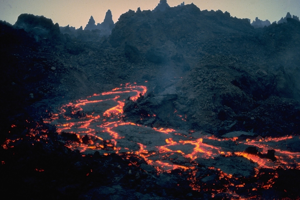 An active incandescent lava channel appears on the spine-covered surface of a rugged aa lava flow at Nyamuragira volcano on January 7, 1982.   During this eruption, which began on December 25, 1981, a fissure on the SE flank produced a lava flow that within a few days was 400-m wide.  It traveled 26 km through forests on the volcano's NE flanks.  This was the longest historical lava flow from Nyamuragira.  Substantial tephra emission during the eruption damaged vegetation.  Copyrighted photo by Katia and Maurice Krafft, 1982.