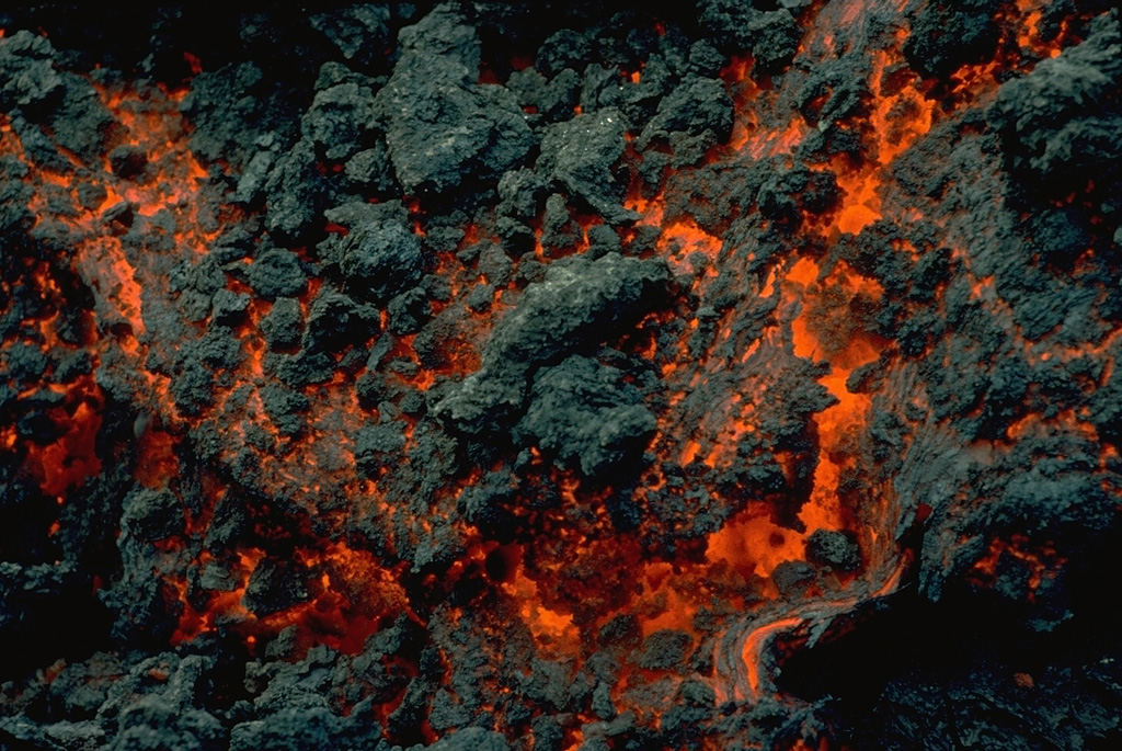 Incandescent lava is visible in the advancing front of an aa lava flow from Nyamuragira on 8 January 1982.  The dark irregular areas are already-solidifed masses that can be carried along by the flow and then break off, forming the rough, clinkery surface of aa flows.  The December 1981 to January 1982 eruption produced the volcano's longest historical lava flow, which traveled 26 km from its SE-flank vent. Copyrighted photo by Katia and Maurice Krafft, 1982.