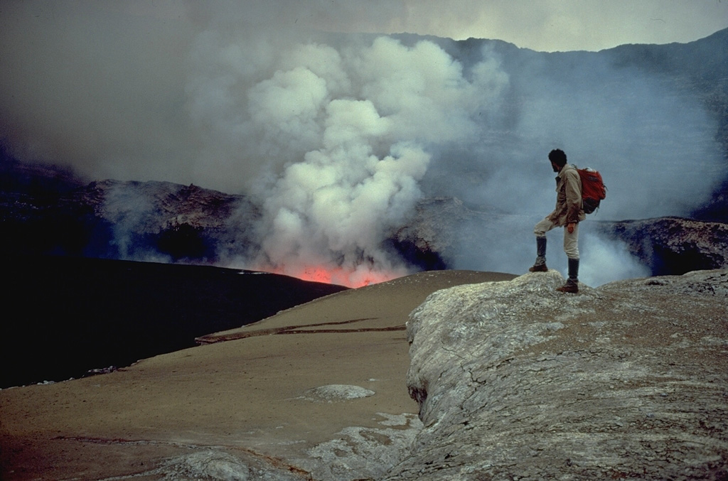 Smoke rises above a lava fountain in the lava lake at the summit of Nyiragongo volcano in June 1973.  Long-term lava lake activity had been occurring since at least 1927.  The observer is standing on one of several terraces left by collapse of solidified lava lakes at higher levels.  Rapid fluctations of the level of the lava lake have occurred since it was first scientifically documented in the 1950's.  The century-long lava lake drained suddenly in 1977. Copyrighted photo by Katia and Maurice Krafft, 1973.