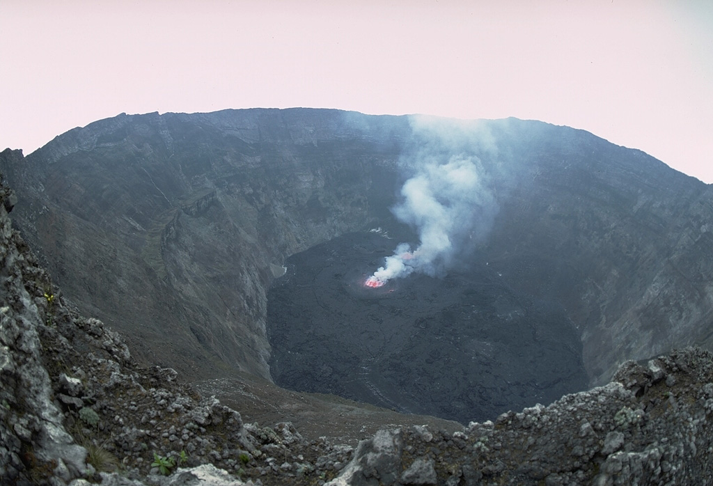 One of Africa's most notable volcanoes, Nyiragongo contained a lava lake in its deep summit crater that was active for half a century before draining suddenly in 1977.  This July 23, 1982 photo from the south crater rim shows a small active lava lake surrounded by black lava flows from the new June 1982 lava lake.  Narrow benches halfway up the steep-walled, 1.2-km-wide summit crater mark former lava lake levels.  Copyrighted photo by Katia and Maurice Krafft, 1982.