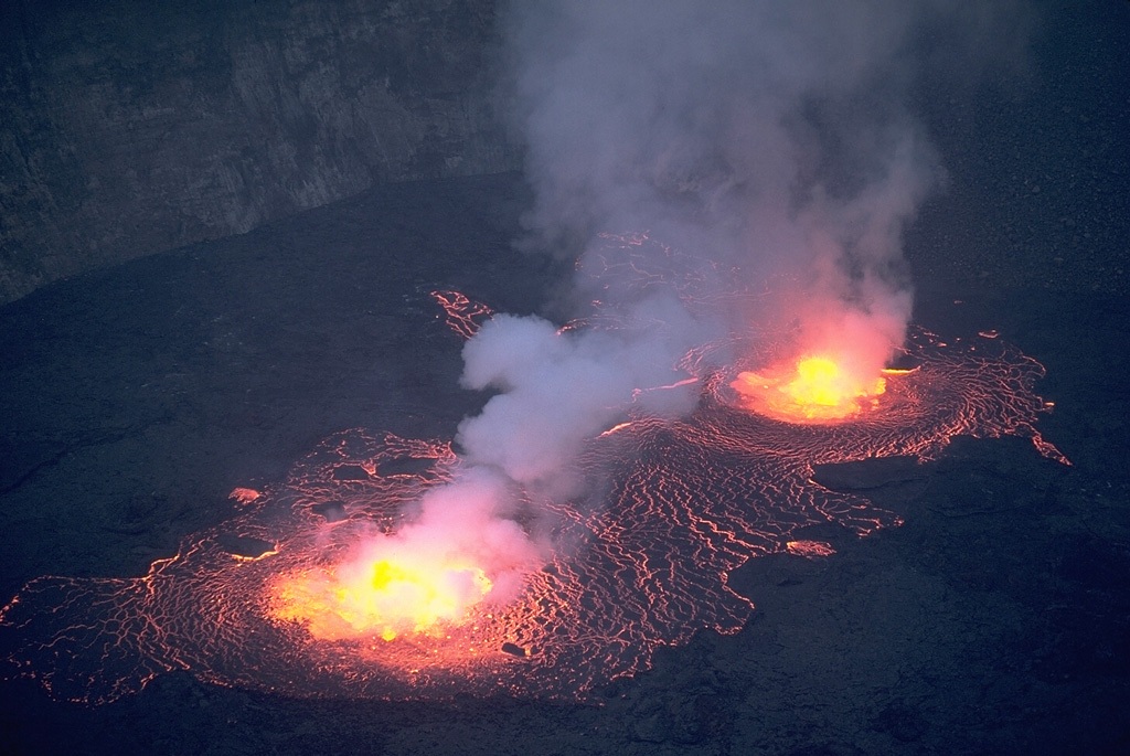Incandescent lava flows spill across the crater floor from two fountains in an active lava lake on the crater floor of Nyiragongo on 23 July 1982.  The lava lake had reappeared on 21 June for the first time since draining suddenly in 1977, leaving a steep-walled, 800-m-deep crater.  Nyiragongo is renowned for its long-term lava lakes.  Prior to 1977, a lava lake had persisted in the 1.2-km-wide crater for half a century.  By the time of this photo, a month after its return to the crater, the lava lake had risen 300 m.  It rose another 100 m by 15 September. Copyrighted photo by Katia and Maurice Krafft, 1982.