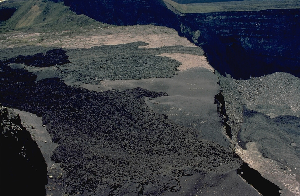 The west rim of the summit caldera provides a view of two lava flows that traveled across a bench on the caldera floor.  The darker flow in the foreground was produced during an eruption in 1972.  It partially overrode the gray-colored flow beyond it produced in a 1965 eruption.  Both flows traveled across the broad bench, one of four large platforms within the caldera, before dropping to the lower floor of the Choungou-Chahale caldera.  The morphology of the compound, 3 x 4 km summit caldera has frequently been modified during historical eruptions. Copyrighted photo by Katia and Maurice Krafft, 1980.
