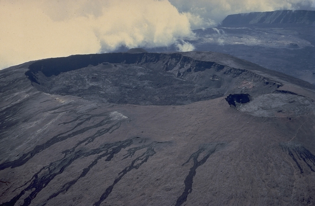 Oval-shaped Dolomieu crater, seen here in a 1973 aerial view from the NE, truncates the summit of a large lava shield that has grown in the summit caldera of Piton de la Fournaise volcano.  The southern wall of the 8-km-wide caldera is visible at the upper right.  The eastern rim of the smaller Bory crater at the right center is cut by Dolomieu crater.  The unvegetated lava flows in the foreground originated in historical time from circumferential fissures near the summit of the lava shield. Copyrighted photo by Katia and Maurice Krafft, 1973.