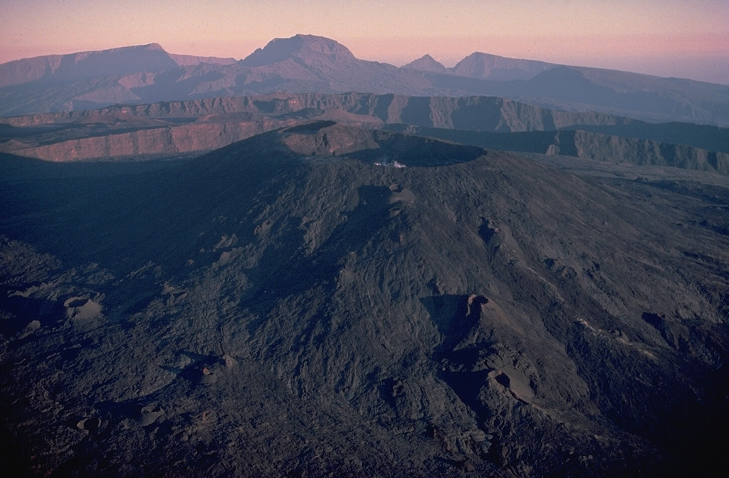 Dolomieu crater (center) caps the summit lava shield of Piton de la Fournaise volcano.  This 1987 aerial view shows the scarps of two large calderas formed by eastward landsliding of the summit of the volcano.  The more distant caldera formed about 65,000 years ago, and the closer one, inside which the modern lava shield was constructed, originated less than 5000 years ago.  Unvegetated fissure-fed lava flows mantle the flanks of the lava shield.  The deeply dissected Pleistocene Piton des Neiges shield volcano forms the western horizon. Copyrighted photo by Katia and Maurice Krafft, 1987.