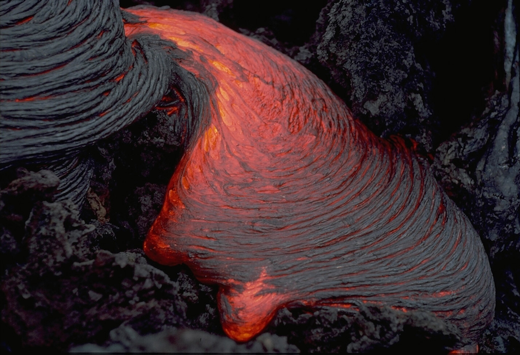 A lobe of ropy pahoehoe lava slowly oozes across the surface of a lava flow at Piton de la Fournaise volcano in the Indian Ocean in March 1976.  The wrinkled surface of pahoehoe lava forms when the still molten interior advances, dragging along the solidifying crust of the flow.  Areas of incandescent lava can be seen between the surface coils of the more solidifed lobe at the upper left.   Copyrighted photo by Katia and Maurice Krafft, 1976.