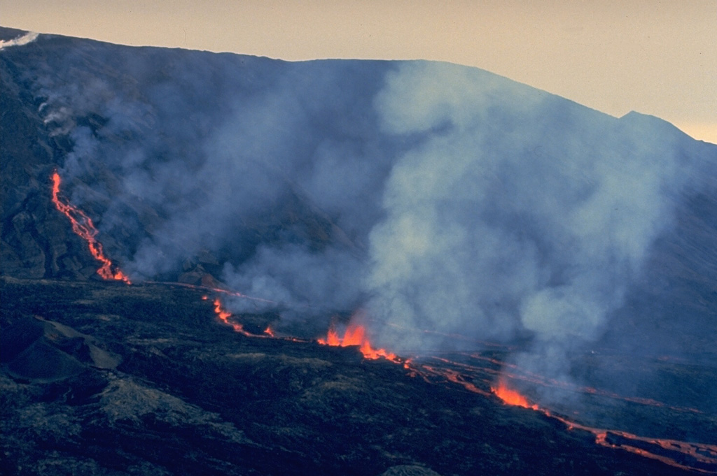 Lava fountains rise along an eruptive fissure cutting the north flank of Dolomieu crater on Piton de la Fournaise in August 1985, during one of the early stages of an eruption that lasted until 1988.  Steam clouds at the upper left mark the higher part of the eruptive fissure, which extends radially away from the crater rim.  Incandescent lava flows fed from the fissure are visible at the left center and behind the steam clouds. Copyrighted photo by Roland Benard, 1985 (courtesy of Katia and Maurice Krafft).