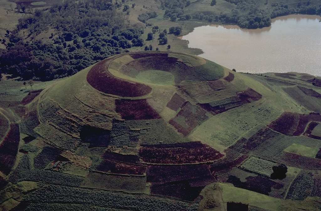 The Itasy volcanic field in central Madagascar contains numerous scoria cones, lava domes, and maar craters.  The entire surface of this scoria cone, which has a shallow summit crater that is breached to the right, is mantled by geometric croplands of local farmers. Copyrighted photo by Katia and Maurice Krafft, 1973.