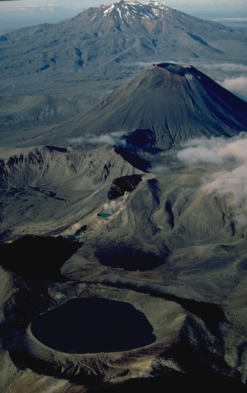 An aerial view from the north shows the Tongariro volcanic centre in the foreground with massive Ruapehu volcano in the background.  The Tongariro volcanic centre consists of a dozen or more partially overlapping volcanoes.  The 400-m-wide Blue Lake crater, in the foreground, erupted about 9700 years ago.  The small Red Crater, at the center, had minor eruptions during the 19th and 20th centuries, and the symmetrical Ngauruhoe stratovolcano behind it has been one of New Zealand's most frequently active volcanoes during historical time. Copyrighted photo by Katia and Maurice Krafft, 1986.