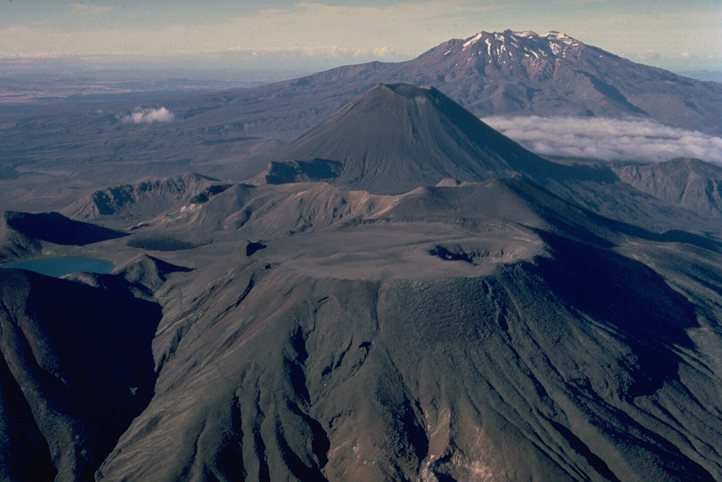 The Tongariro volcanic massif is a complex of more than a dozen composite volcanic cones erupted along a NE-SW line.  Flat-topped North Crater in the foreground and conical Ngauruhoe behind it are the two most prominent Tongariro volcanoes.  North Crater's broad, flat top is the surface of a former lava lake; a small explosion crater later punctured its right side.  Most historical eruptions of Tongariro have originated from the symmetrical Ngaurohoe stratovolcano.  Ruapehu volcano forms the broad massif in the distance to the south. Copyrighted photo by Katia and Maurice Krafft, 1986.