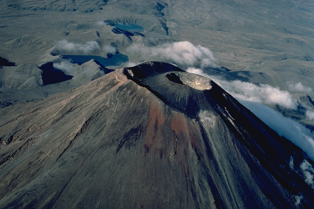 Symmetrical Ngauruhoe volcano, seen here in an aerial view from the NE, contains a compound summit crater.  The most recent eruptions have occurred from the crater of a small cinder cone that was constructed within the outer summit crater beginning in 1954.  Ngauruhoe is one of New Zealand's most active volcanoes; frequent changes in the morphology of the summit crater have been documented during historical eruptions.  The two lakes in the background are the Tama Lakes, flank vents of Ruapehu volcano. Copyrighted photo by Katia and Maurice Krafft, 1986.