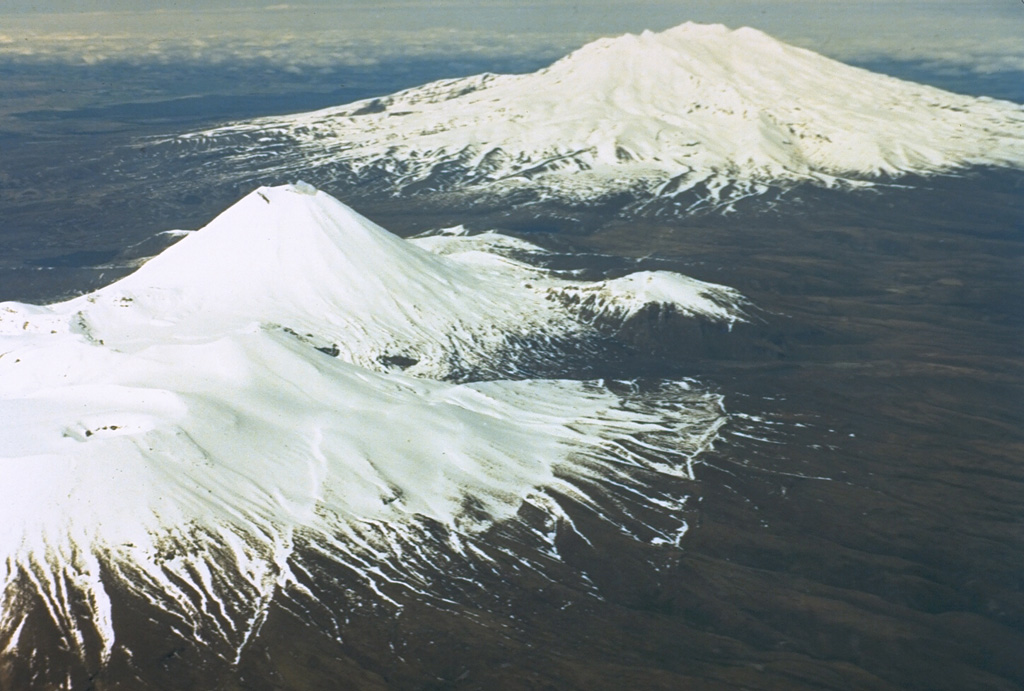 Tongariro, in the foreground, is a cluster of about a dozen cones and craters dominated by the symmetrical Ngauruhoe cone. Ruapehu, in the background to the south, contains a frequently active crater lake. The two volcanoes have been two of the most frequently active in New Zealand. Photo by New Zealand Geological Survey, 1981.