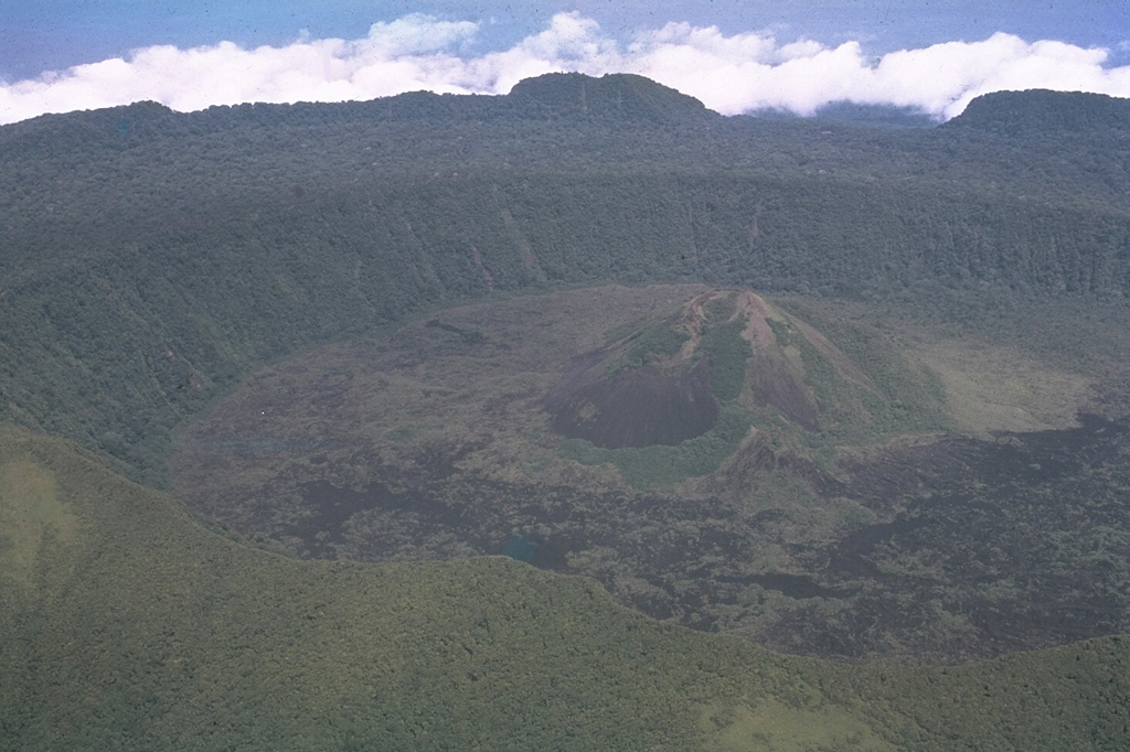 Two nested calderas are visible in this aerial view from the E of the Karkar summit, taken before the 1974 eruption. The 5.5-km-wide outer caldera formed during one or more eruptions, the last of which occurred 9,000 years ago, and the rim forms the horizon. The 3.2-km-wide inner caldera was formed sometime between 1,500 and 800 years ago and its floor is covered by young lava flows. Bagiai cone (right center) has been the source of most eruptions dating back to 1643. Photo by Wally Johnson, 1974 (Australia Bureau of Mineral Resources).