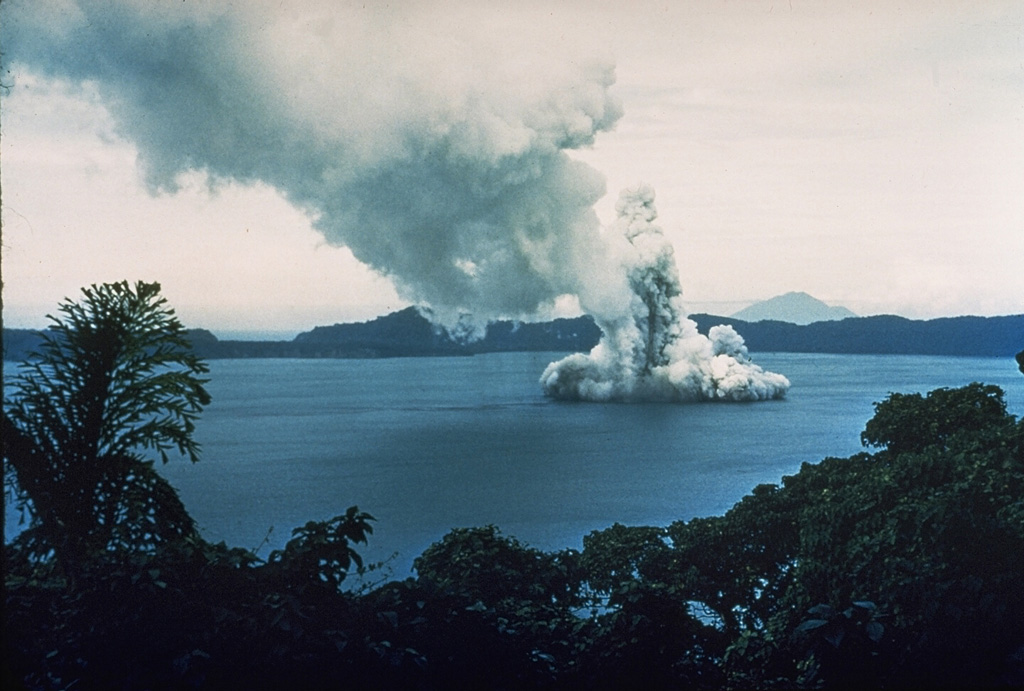 Phreatic explosions from Motmot Island in Lake Wisdom, on Long Island, during a 1953-54 eruption. The Surtseyan explosion produced a jet of steam and ash seen rising above the lake, and a base surge traveling radially away from the vent across the surface. The western wall of the 10 x 12.5 km Long Island caldera is 200-300 m high in the distance. Twentieth century eruptions at Long Island have originated from vents at or near Motmot Island. Photo by John Best (courtesy of Wally Johnson, Australia Bureau of Mineral Resources).