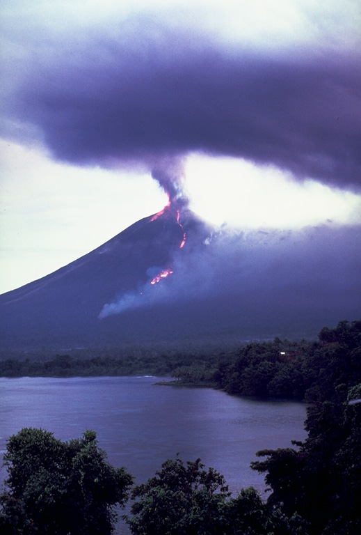 An ash plume erupts from the summit crater of Ulawun volcano on 21 November 1985. Smoke from burning vegetation rises from the front of an incandescent lava flow that is descending the NW flank. Strombolian eruptions had begun four days earlier and lava emission began the following day. The lava flow front bifurcated on the lower flanks of the volcano and the eastern lobe traveled slightly farther, eventually reaching 5.5 km from the summit. Photo by Wally Johnson, 1985 (Australia Bureau of Mineral Resources).