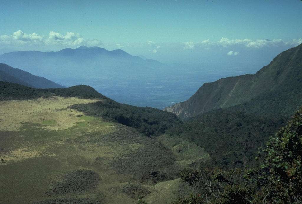 The flat area to the left is Alun-Alun, the uppermost of four large craters on Papandayan volcano. The v-shaped valley in the center of the photo extending to the NE is the breach left by collapse of Papandayan volcano in 1772. The volcano in the distance on the left horizon is Gunung Guntur, another historically active volcano bordering the Garut Plain. Photo by Tom Casadevall, 1986 (U.S. Geological Survey).