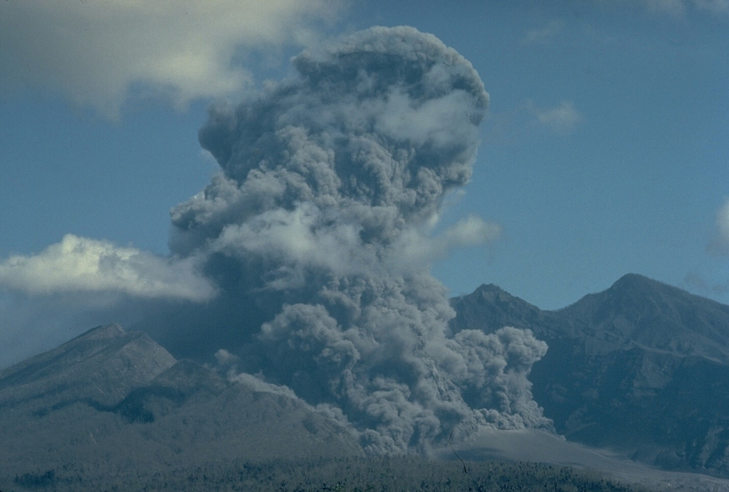 An eruption plume rises above the vent of Galunggung volcano in September 1982, and at the lower right ash billows above a pyroclastic flow descending the floor of the breached crater of Galunggung.  Intermittent eruptions sometimes accompanied by pyroclastic flows and lahars had occurred since April 5, 1982. Copyrighted photo by Katia and Maurice Krafft, 1982.