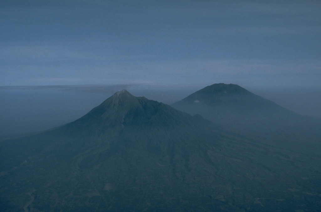 Merapi (left) and Merbabu (right) volcanoes tower above the lowlands between the cities of Yogyakarta and Surakarta in this aerial view from the SE. Both have erupted during historical time, but Merapi is by far the more vigorous of the two. Activity at Merapi frequently includes lava dome and collapse that produces pyroclastic flows and lahars that travel down the valleys, often with catastrophic results. Photo by Jeff Post, 1991 (Smithsonian Institution).