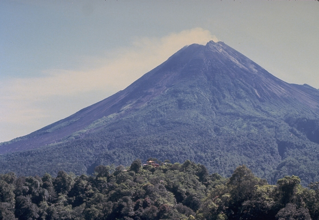Merapi volcano, one of Indonesia's most active, rises above the city of Yogyakarta in central Java. This May 1987 view from the southern flank with the red-roofed Plawangan observatory on the forested ridge in the foreground shows Merapi's degassing summit lava dome. Unvegetated areas on the left result from periodic rockfalls, avalanches, and pyroclastic flows (block-and-ash flows) from the growing lava dome. Photo by Tom Casadevall, 1986 (U.S. Geological Survey).