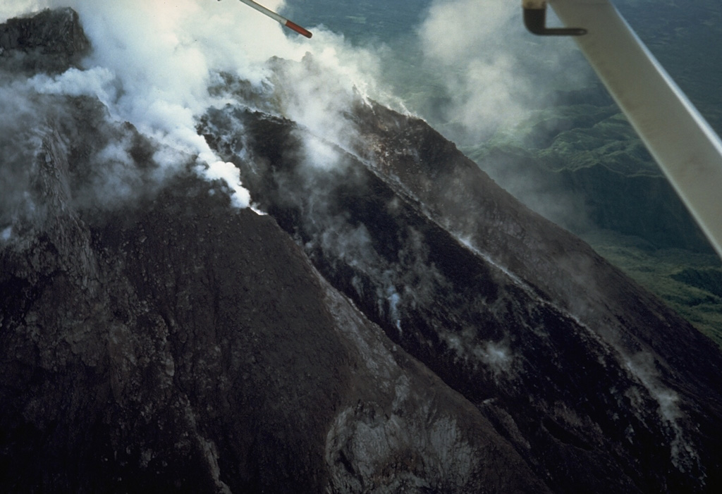An aerial view from the WSW on 6 January 1987 shows a black lava flow descending from the summit crater of Merapi. During a five-day period beginning 10 October 1986, the dome was partially destroyed by a series of dome-collapse pyroclastic flows (block-and-ash flows). The period of dome growth and lava effusion seen in this photo then began and continued for several years before stabilizing in mid-1990. Photo by Tom Casadevall, 1987 (U.S. Geological Survey).