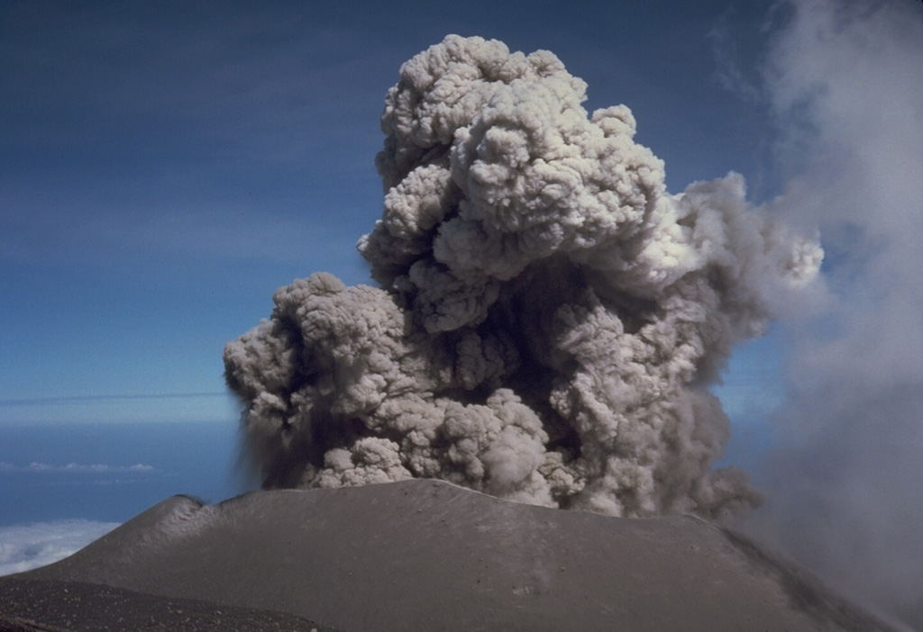 An eruption plume rises above the summit crater of Semeru on 17 August 1985. Semeru has been in near-continuous activity since 1818, producing frequent small-to-moderate explosions that eject ash and volcanic bombs. Some larger eruptions have produced pyroclastic flows and lahars that have reached the foot of the volcano. Photo by Tom Casadevall, 1985 (U.S. Geological Survey).