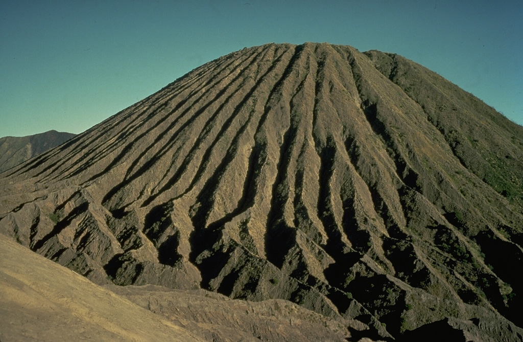 Erosion of friable tephra deposits forming the Batok pyroclastic cone on the floor of Tengger caldera has created dramatic erosional furrows that reach from the summit to the base of the cone.  Batok is one of the youngest of the post-caldera cones; charcoal from a scoria layer on the eastern flank of Batok was radiocarbon dated at about 360 years ago.  Only the historically active cone of Bromo, from where this photo was taken, is younger. Copyrighted photo by Katia and Maurice Krafft, 1976.