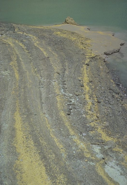 The yellow stripes in the foreground are strandlines of elemental sulfur particles that have been washed up on the shoreline of Kawah Ijen lake by wave action. The crater walls of Kawah Ijen have been the site of a long-term sulfur-mining operation. Photo by Tom Casadevall, 1987 (U.S. Geological Survey).