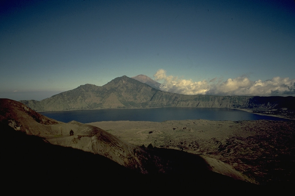 The SE wall of Batur's 10 x 13 km outer caldera rises above the caldera lake, as seen from Batur volcano, which grew within a 7.5-km-wide, inner caldera.  Lava flows from 19th century eruptions of Batur volcano, on the island of Bali, form the lake's near shoreline, which is near the margin of the buried eastern rim of the inner caldera.  Agung volcano, at the eastern tip of Bali, is visible in the distance. Copyrighted photo by Katia and Maurice Krafft, 1971.