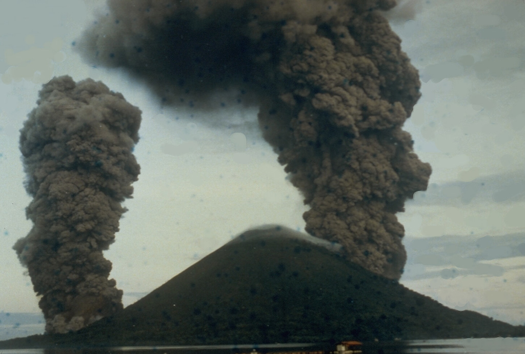 This dramatic photo taken from the SE on May 9, 1988, shows ash-rich eruption plumes rising from vents on the north and south sides of the island.  The blue dots are a result of film damage.  The eruption occurred from a 3-km-long arcuate fissure that cut the island from the south coast to the 200-m elevation on the north flank.  The eruption plume reached a maximum height of 16.5 km.  Lava flows reached the north, NW, and south coasts, destroying two villages. Photo by I. Yoshida, 1988 (courtesy of Volcanological Survey of Indonesia).