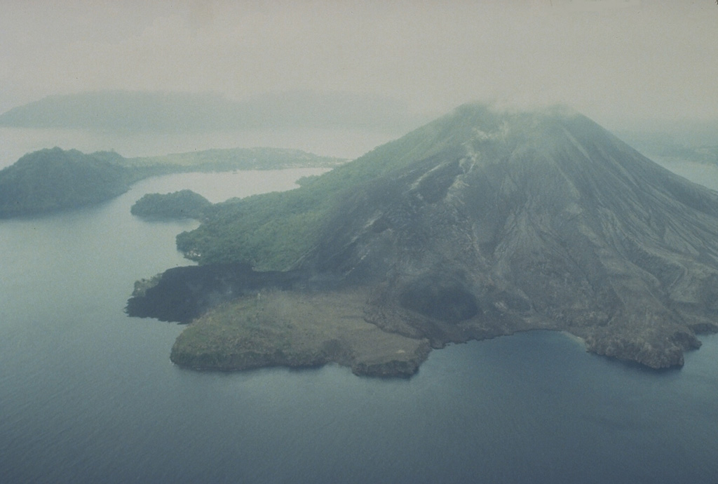 The May 1988 north-flank lava flows reached the sea at two locations.  The flows originated from a fissure that can be seen descending diagonally to the left from the summit.  The Pasir Besar lava flow was erupted from a vent at about 300-350 m elevation and reached the sea along almost the full length of Pasir Besar bay at the right-center.  The Batu Angus lava flow originated from a vent at 200 m elevation and reached the sea at the left-center after overrunning houses in the villages of Kalobi and Batu Angus. Photo by Tom Casadevall, 1988 (U.S. Geological Survey).