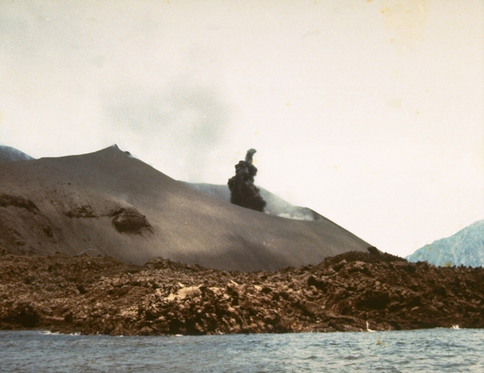 A small dark ash plume begins rising from the crater of Anak Krakatau in October 1978. Explosive activity began in July and lasted until November. A lava flow from a previous eruption forms the coast in the foreground. Photo by J. Matahelumual, 1978 (Volcanological Survey of Indonesia).