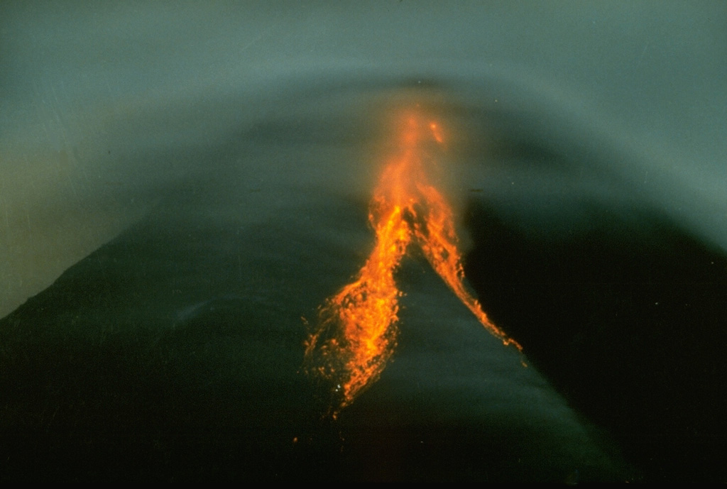 Incandescent rockfalls can be seen accompanying growth of a Merapi lava dome beneath clouds covering the summit in this 1993 nighttime view. Periodic collapse of Merapi's lava dome has produced pyroclastic flows down the western and southern flanks that have devastated populated areas and agricultural lands. Photo by Ruska Hadian, 1993 (Volcanological Survey of Indonesia).