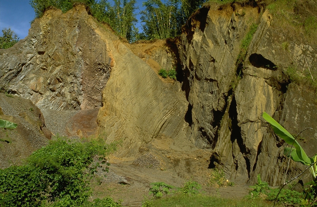 An outcrop in a quarry showing the internal structure of a debris avalanche hummock, with bedded layers of tephra on the right and a segment of a lava flow on the left. Both the massive lava and the unconsolidated tephra layers were transported relatively intact for about 30 km within a debris avalanche from Raung volcano in eastern Java. The preservation of original stratigraphy from within the volcano is a common feature of debris avalanche deposits. Photo by Lee Siebert, 1995 (Smithsonian Institution).