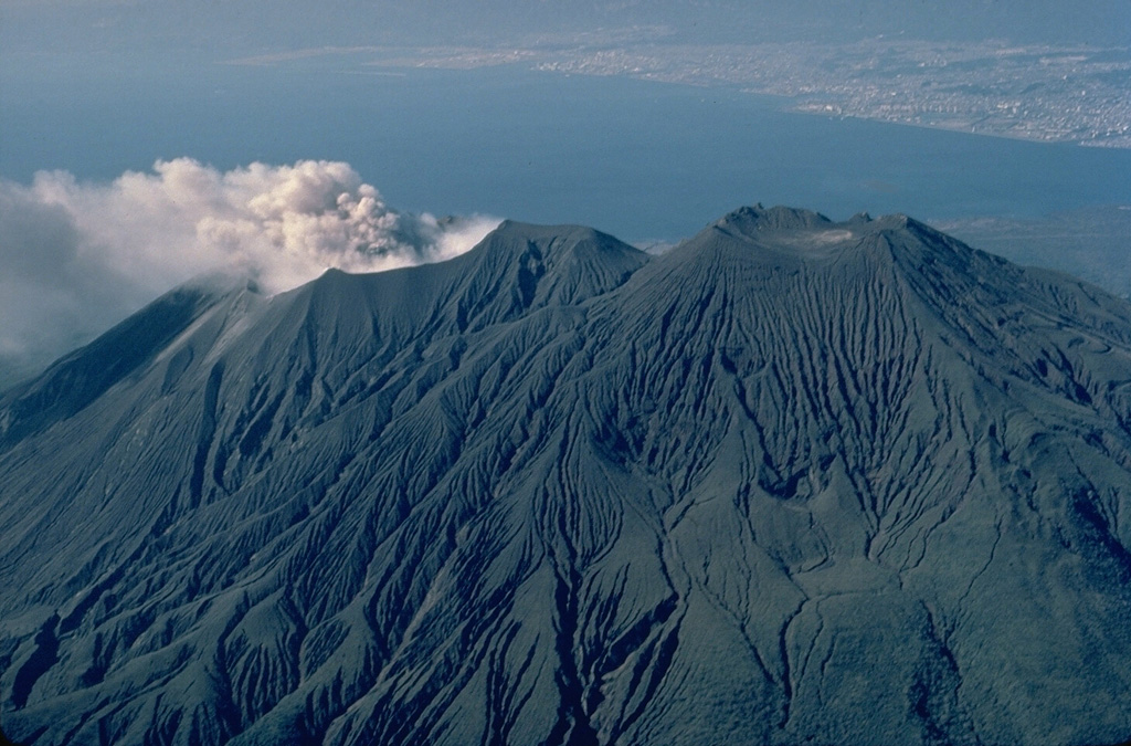 An aerial view from the NE shows the three summit cones of Sakura-jima.  Kita-dake, the northern cone, is at the right, Naka-dake in the center, and a plume of ash and steam rises from Minami-dake, the historically active southern crater.  The city of Kagoshima appears at the upper right across Kagoshima Bay. Copyrighted photo by Katia and Maurice Krafft, 1981.