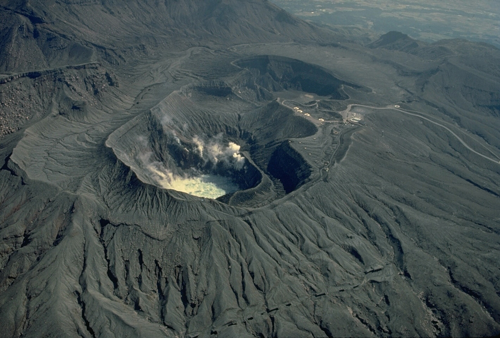 A group of overlapping craters marks the summit of Naka-dake volcano one of Japan's most active volcanoes.  Seen here from the north, steam rises from a hot lake in the near crater and from fumaroles along its wall.  Frequent explosive eruptions deposit ash and blocks on the flanks of the cone, leaving it unvegetated.  A parking lot can be seen on the west rim of the crater, which is one of Kyushu's most popular tourist destinations. Copyrighted photo by Katia and Maurice Krafft, 1981.