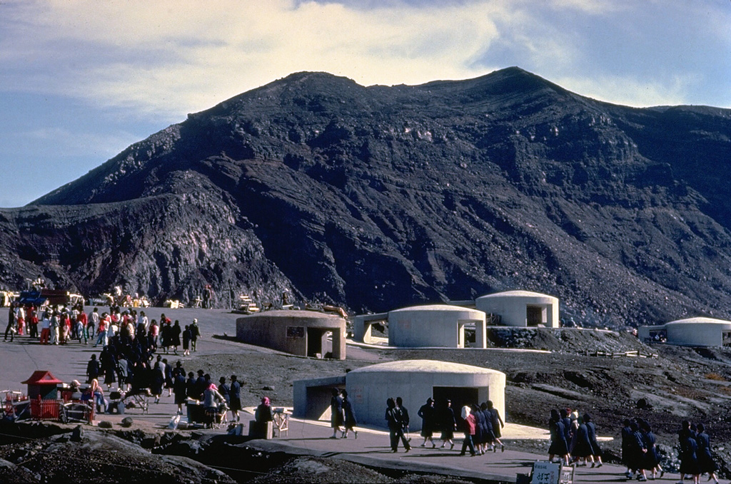 Tourists pack a parking lot at the summit crater of Naka-dake, the historically active crater of Aso volcano.  The crater rim is readily accessible by toll road or aerial cable car and is a popular tourist destination.  The circular concrete structures are emergency shelters to protect tourists in the event of a sudden volcanic eruption.  Frequent, but intermittent, explosive eruptions eject ash and blocks onto the flanks of the cone and often prompt closure of the summit to visitors. Copyrighted photo by Katia and Maurice Krafft, 1981.