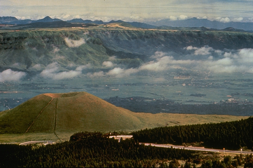 Kometsuka scoria cone (lower left) formed about 1,800 years ago in the Aso caldera complex. Clouds are above the broad caldera floor surrounding the cone complex and the NW wall of the 24-km-wide caldera is visible behind the cone. The caldera formed during four major Pleistocene explosive eruptions between 300,000 and 80,000 years ago. Photo by Ichio Moriya (Kanazawa University).