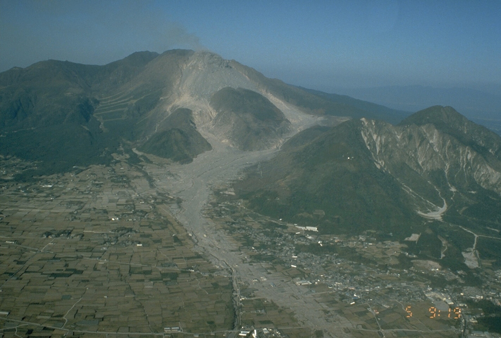 Shichimenzan and Mayuyama are two lava domes that form the forested peaks in this 5 November 1991 view from the SW. Mayuyama, the closest dome, has an E-facing scarp resulting from a major debris avalanche in 1792 that swept into the Ariake Sea. The avalanche produced a catastrophic tsunami that swept the coast of Shimabara Peninsula and traveled across the bay, causing nearly 15,000 fatalities. The light-colored areas in the foreground are 1991 block-and-ash flow deposits. Photo by T. Kobayashi, 1991 (Kagoshima University).