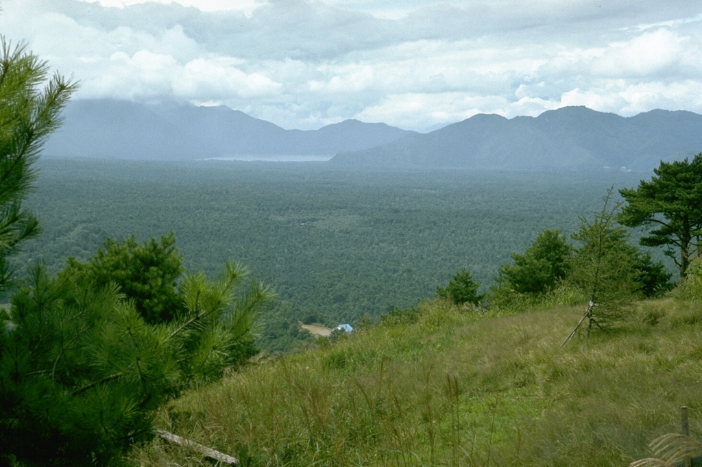 This entire flat forested plain, known as Aokigahara ("Blue Tree Plain") is underlain by a single massive lava flow from Fuji. A major explosive and effusive eruption began in June 864 CE from Nagaoyama, a vent on the NW flank. Lava flowed into lakes Motosu and Senoumi, destroying lakeshore houses. It divided Lake Senoumi into the two present-day lakes of Shojiko(seen here in the distance against a Tertiary mountain range to the NW) and Saiko. The Aokigahara Marubi lava field covered an area of 32 km2. Photo by Lee Siebert, 1977 (Smithsonian Institution).