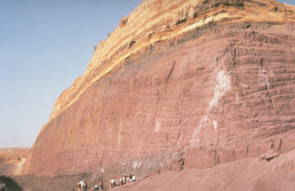 Geologists examine a quarry in a post-caldera cone of the Vulsini volcanic complex. The base of the quarry exposes thick, red-colored scoria deposits produced by Strombolian eruptions that built a scoria cone. These are overlain by bedded, yellowish pyroclastic surge deposits. Photo by Richard Waitt, 1985 (U.S. Geological Survey).