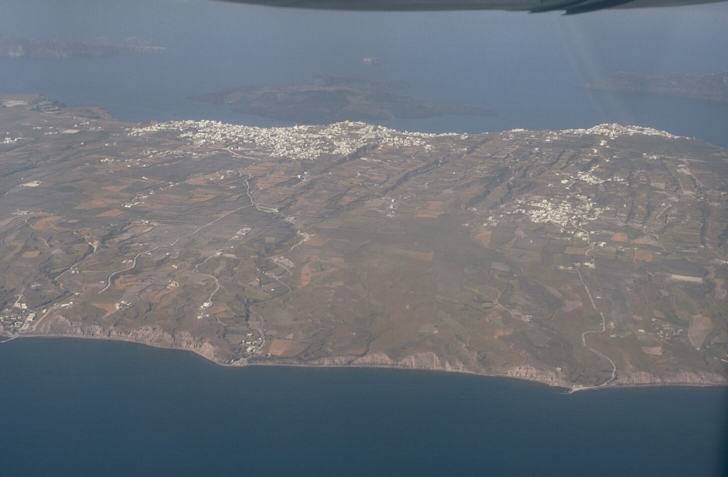 An aerial view from the east overlooks the outer flanks of Santorini's 7.5 x 11 km caldera. The far caldera rim, part of which is flooded by the sea, appears at the upper left and right. The towns of Firá and Merovígli sit on the caldera rim and farms drape the outer flanks of the volcano, which are underlain by deposits from the cataclysmic Minoan-age eruption about 3,500 years ago. The post-caldera islands of Nea and Palaea Kameni (upper-center) were constructed in the middle of the caldera during eruptions dating back to 197 BCE. Photo by Lee Siebert, 1994 (Smithsonian Institution)