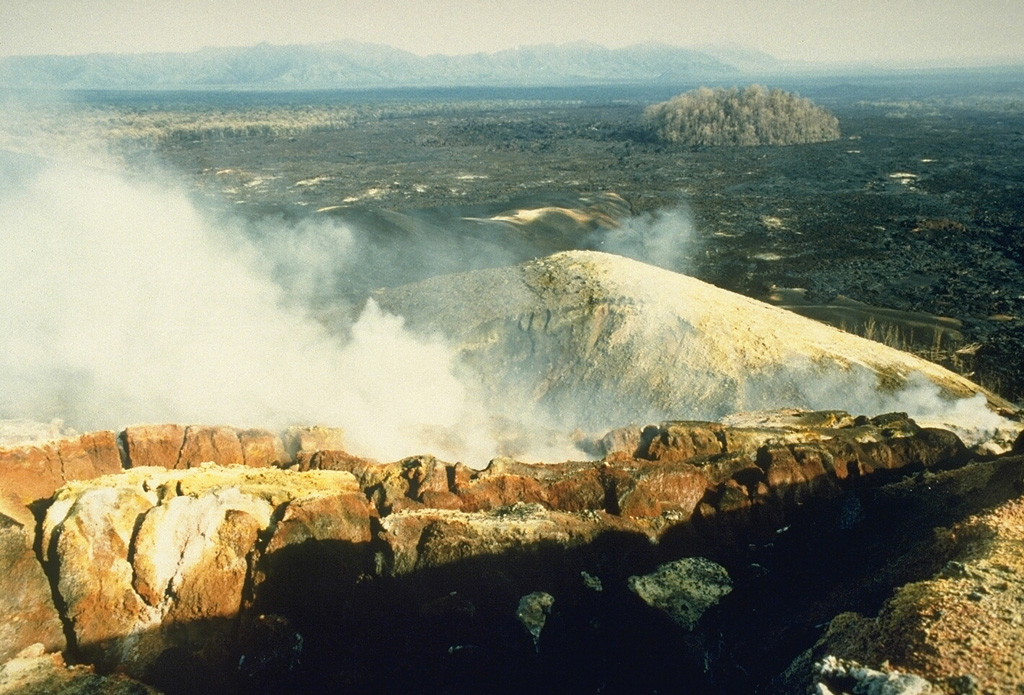 Steam rises from Kimanura scoria cone, which was formed during an eruption from April to August 1989 on the E flank of Nyamuragira. This December 1989 photo looks N across a black lava flow that had traveled about 20 km down the N flank. The flow diverted around a forested "kipuka" of an older cone (upper right), scorching leaves but not burning the trees. The Kimanura vent was the lowest of three that were active in 1989. A small fissure in the summit caldera and another on the upper SE flank also produced lava flows. Photo by Henry-Luc Hody, 1989 (Belgian ambassador).